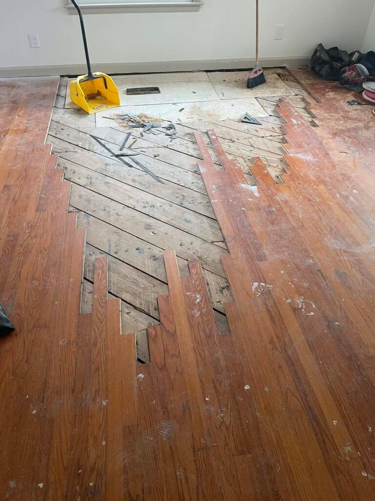 Hardwood floor repair in progress with boards partially removed, a yellow dustpan, a broom, and tools on the floor